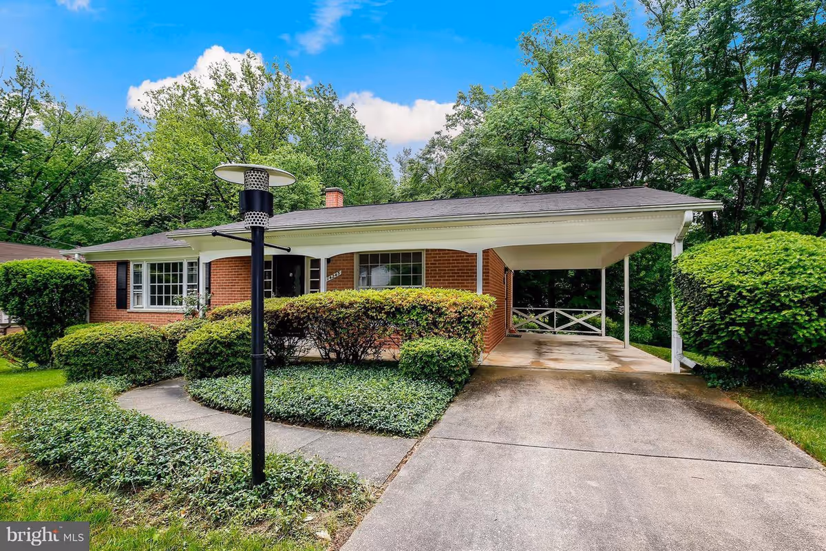 Front exterior view of a single-story brick house with a covered carport, surrounded by green bushes and trees under a blue sky with some clouds.
