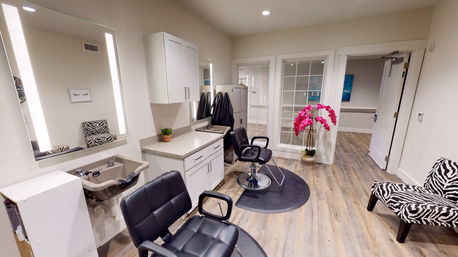 Interior view of a salon area in a senior living facility featuring two black salon chairs, a small sink, a large illuminated mirror, white cabinetry, a potted plant, a zebra-patterned chair, and a vibrant pink orchid plant near a glass-paneled door. The room has light wood flooring and white walls.