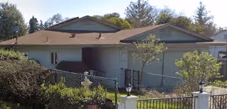 Single-story residential building with a brown roof and light-colored exterior walls, surrounded by trees and bushes, with a chain-link fence in the foreground under a clear sky.