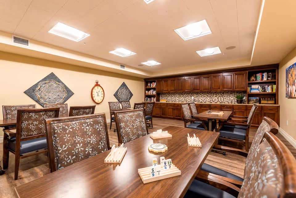 A well-lit room with several wooden tables and cushioned chairs arranged for group activities. The tables have game pieces and racks set up, suggesting a recreational or social space. The back wall features wooden cabinets with a mosaic tile backsplash, shelves with books and games, and decorative wall art including a large clock and two ornate square panels.