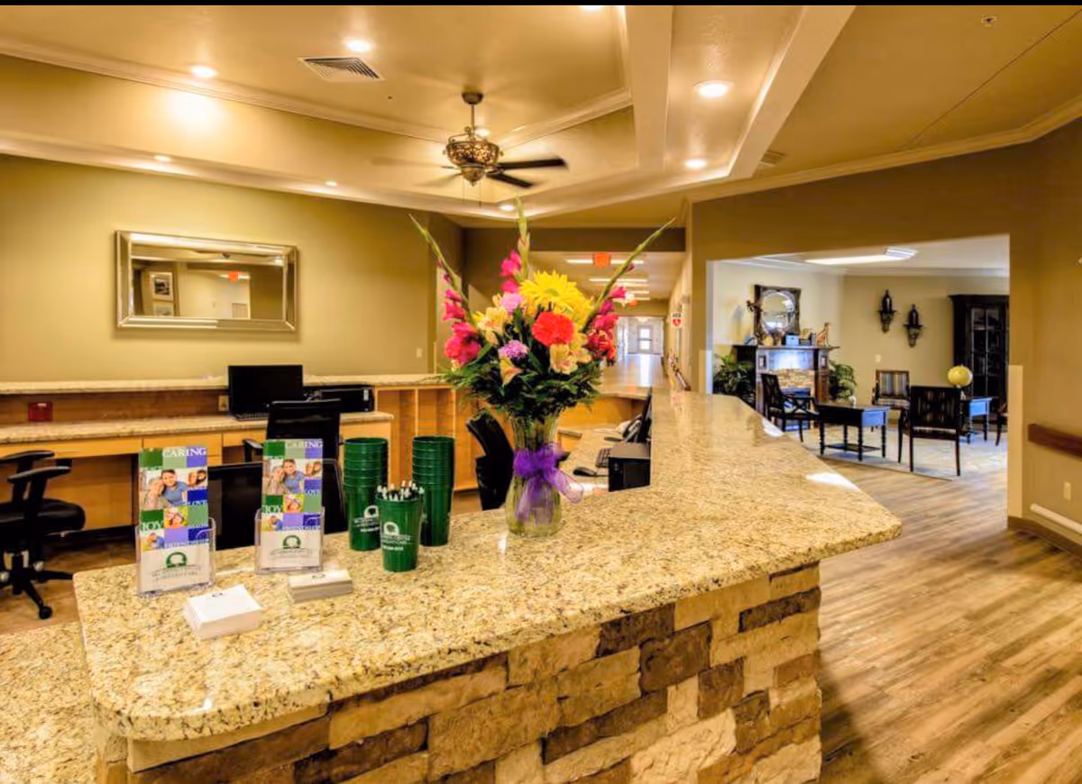 Reception area of Big Spring Center for Skilled Care with a granite countertop desk adorned with a vase of colorful flowers, brochures, and green cups. Behind the desk are office chairs and computer monitors. To the right, there is a seating area with chairs, a table, and decorative wall sconces. The space has warm lighting and wood flooring.