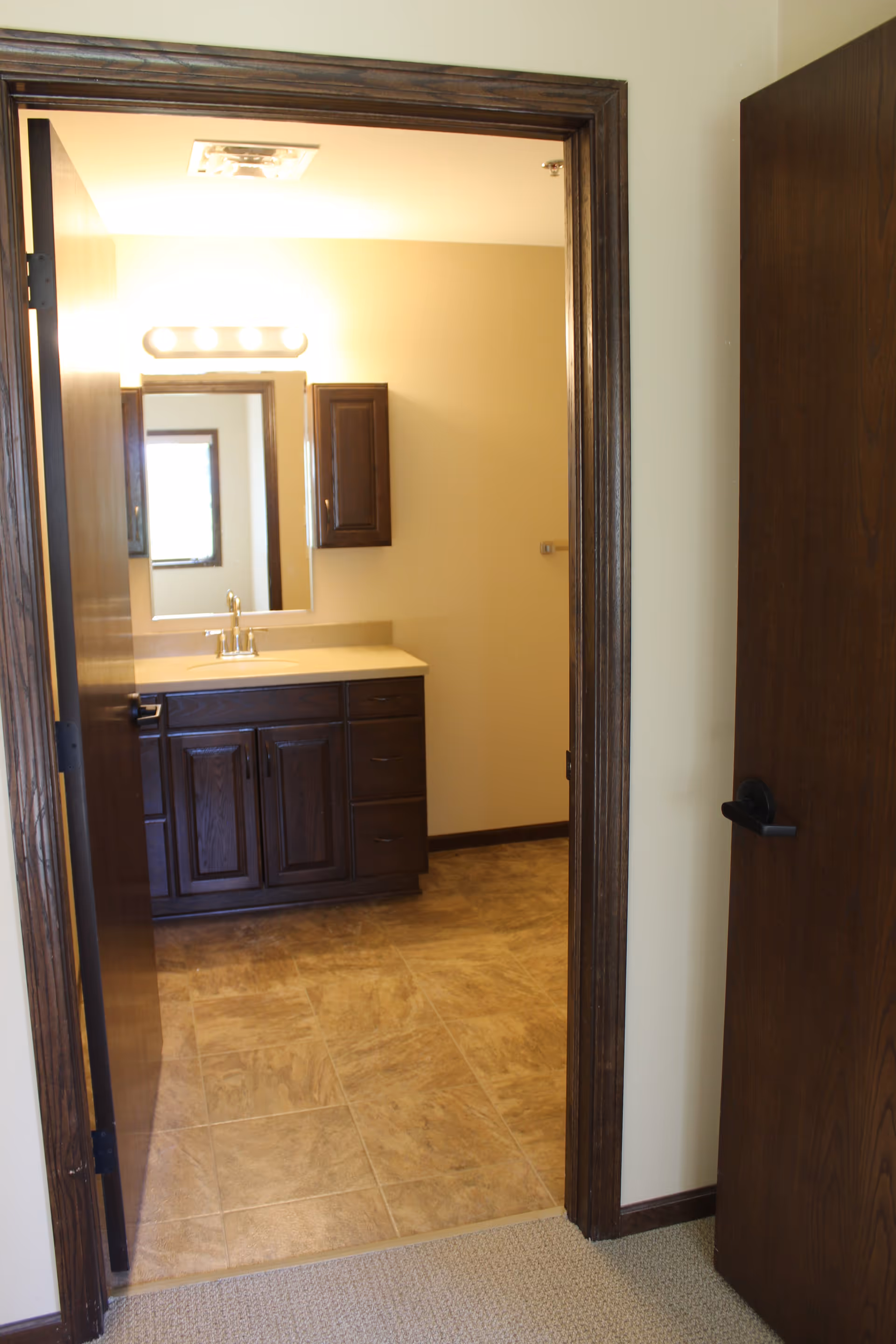 View through an open door into a bathroom with a wooden vanity cabinet, a countertop with a sink and faucet, a wall-mounted cabinet, a mirror, and a light fixture above the mirror. The bathroom floor is tiled, and the walls are painted a light beige color.