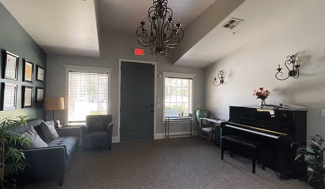 A cozy living room with a dark green door centered between two windows with blinds. To the left, there is a dark gray sofa with pillows, an armchair, a floor lamp, and framed certificates or pictures on a green accent wall. To the right, there is a black upright piano with a bench, a small desk with a chair, wall-mounted candle-style sconces, and some plants. A decorative chandelier hangs from the ceiling.