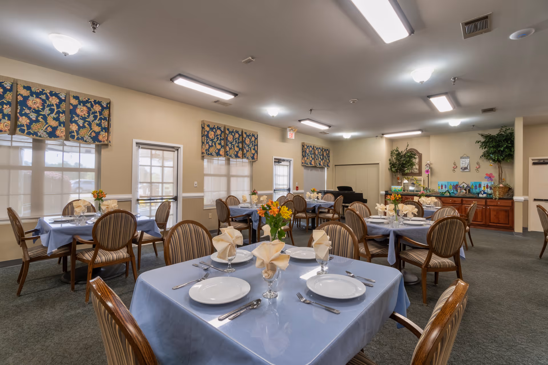 A dining room in an assisted living facility with several tables covered in light blue tablecloths. Each table is set with white plates, silverware, folded beige napkins in glasses, and small floral centerpieces. The room has beige walls, windows with floral valances, and overhead fluorescent lighting. There is a wooden sideboard with decorations and plants in the background.