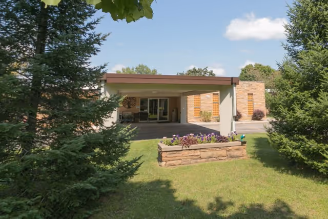 Front exterior view of Greentree Health And Rehab Center showing the entrance with a covered drop-off area, surrounded by green grass, trees, and a flower bed in the foreground under a partly cloudy sky.