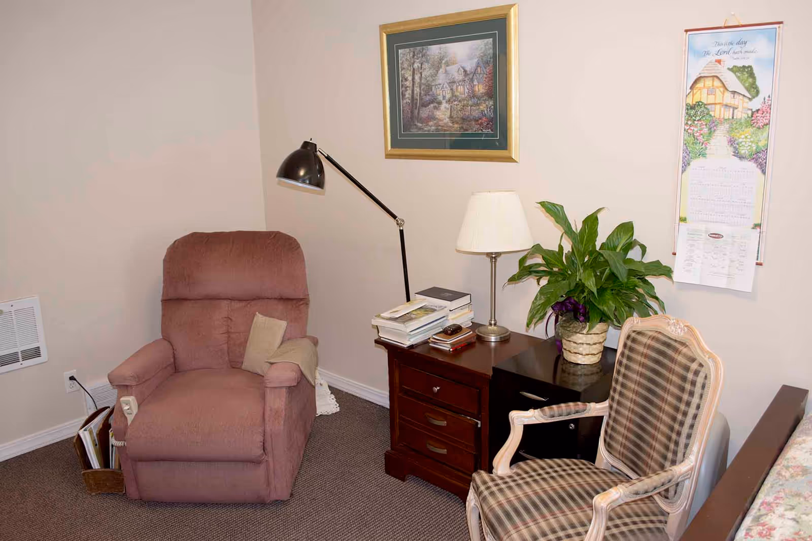 A cozy corner of a room featuring a mauve recliner chair with a beige pillow and blanket, a wooden side table with a stack of books, a table lamp, and a potted plant. Next to the table is a plaid upholstered armchair. The walls are beige with a framed painting of a cottage and a hanging calendar with a house illustration.