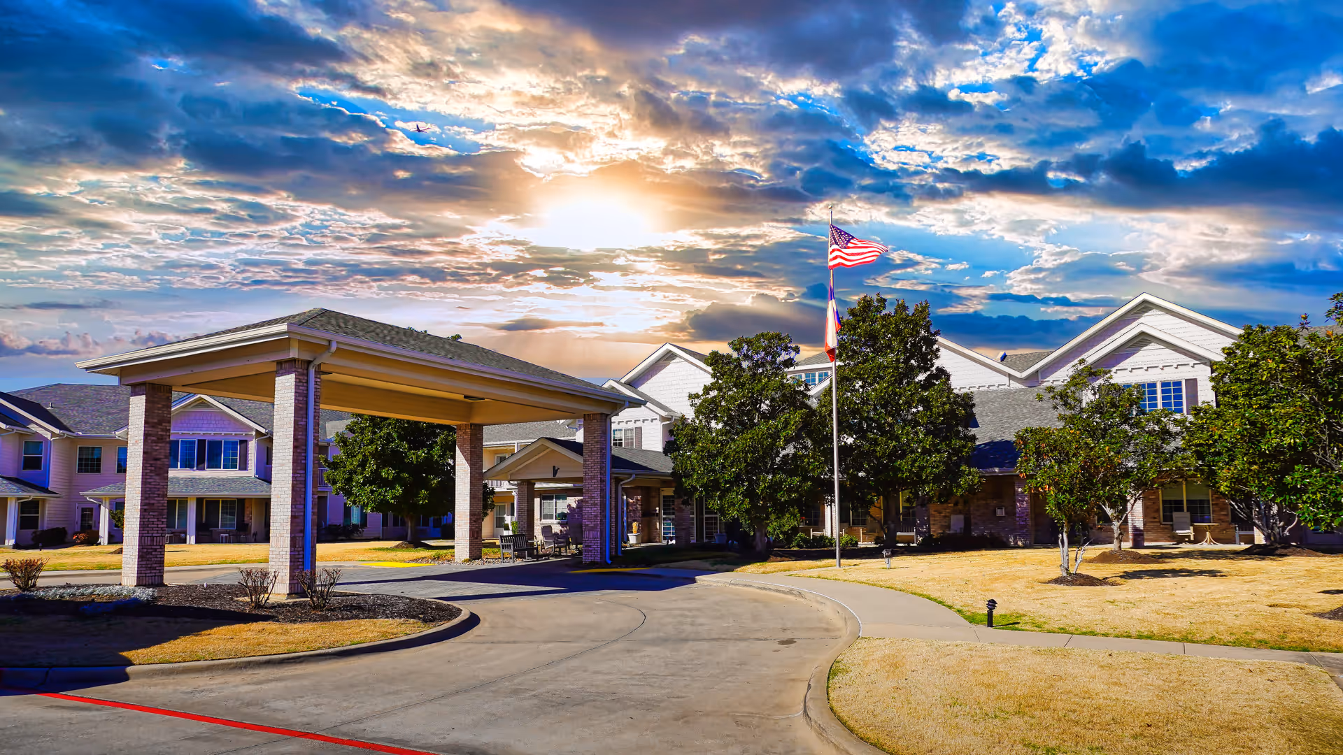 Exterior view of Pinewood Hills - Sky Active Living facility with a covered entrance, a driveway, trees, and an American flag on a flagpole under a partly cloudy sky with the sun shining through.