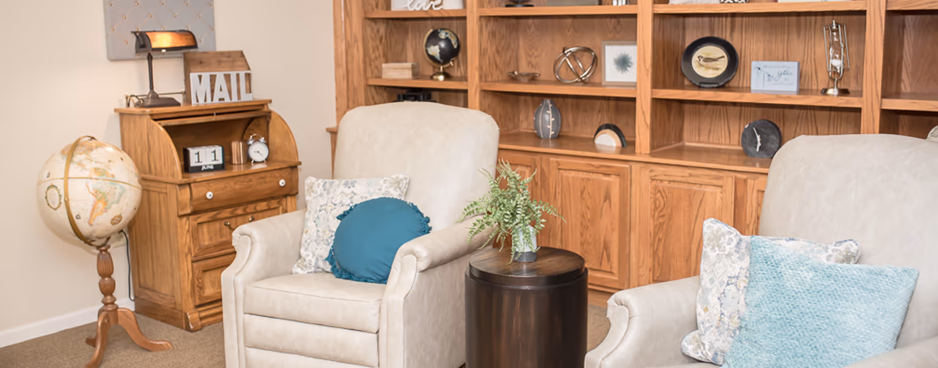 A cozy living room area with two beige armchairs adorned with decorative pillows, a small round wooden side table with a potted plant, a wooden globe stand, and a wooden roll-top desk with a lamp and mail organizer. Behind the chairs is a large wooden built-in bookshelf with various decorative items.