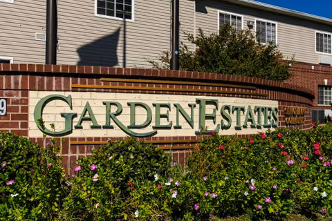Brick and stone sign with green letters reading 'Garden Estates' surrounded by flowering bushes, with a beige residential building in the background under a clear blue sky.