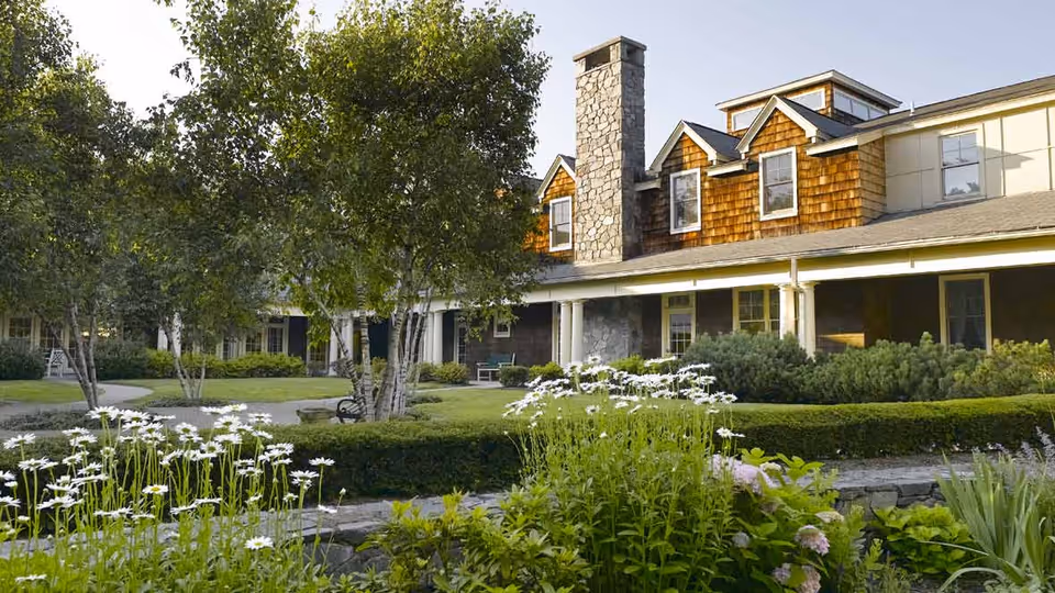 A serene outdoor garden area at Atria Darien featuring lush greenery, white flowers, trees, and a well-maintained lawn in front of a building with a stone chimney and wooden shingles.