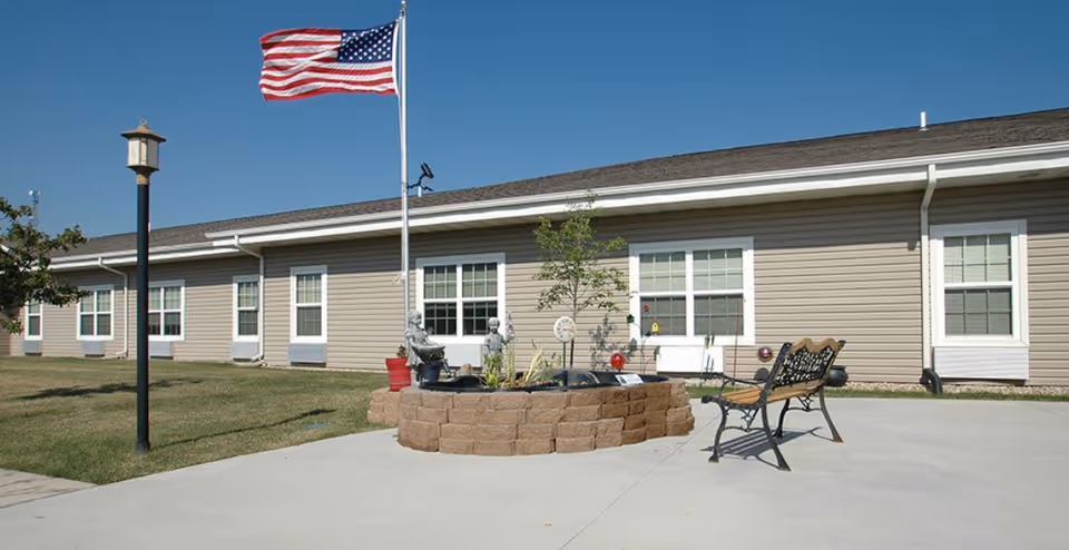 Single-story senior living building front with an American flag, raised planter, and bench on a concrete patio.