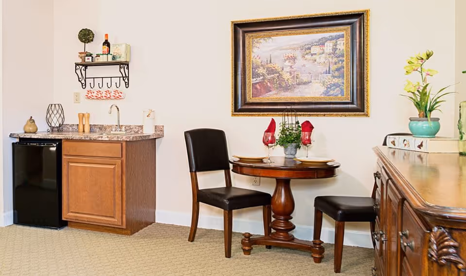 Small dining area with a round wooden table set for two, two chairs, a kitchenette with sink and mini-fridge, and a framed painting on the wall.