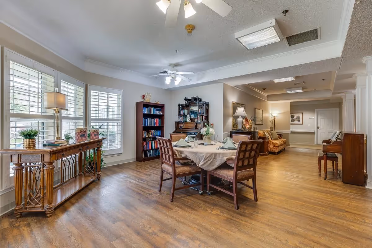 A well-lit common area with wooden flooring featuring a round dining table set with four chairs and place settings. To the left, there is a wooden console table with a lamp and plants in front of large windows with white shutters. In the background, there is a bookshelf filled with books, a decorative cabinet, a sofa, framed artwork on the walls, and a piano on the right side.