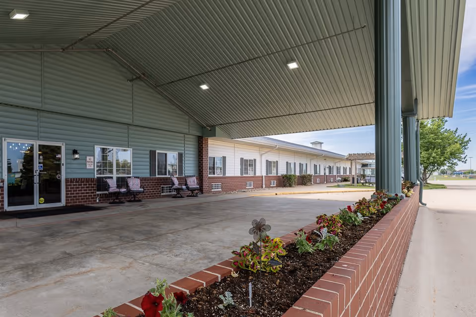 Covered entrance area of Bailey Pointe at Plattsmouth facility with a brick flower bed containing various plants and flowers in the foreground. The building exterior features green siding with brick accents and several windows with black shutters. There are chairs lined up against the wall near the entrance door, and the area is well-lit with ceiling lights under the large roof covering the driveway.