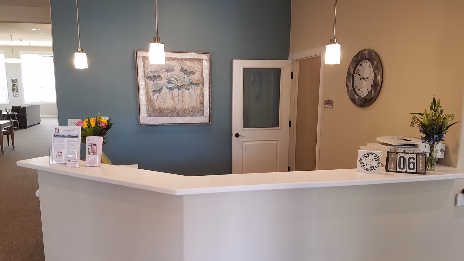 Reception area with a white counter, two pendant lights hanging from the ceiling, a blue accent wall with a floral painting, a door with frosted glass, a wall clock, and flower arrangements on the counter.