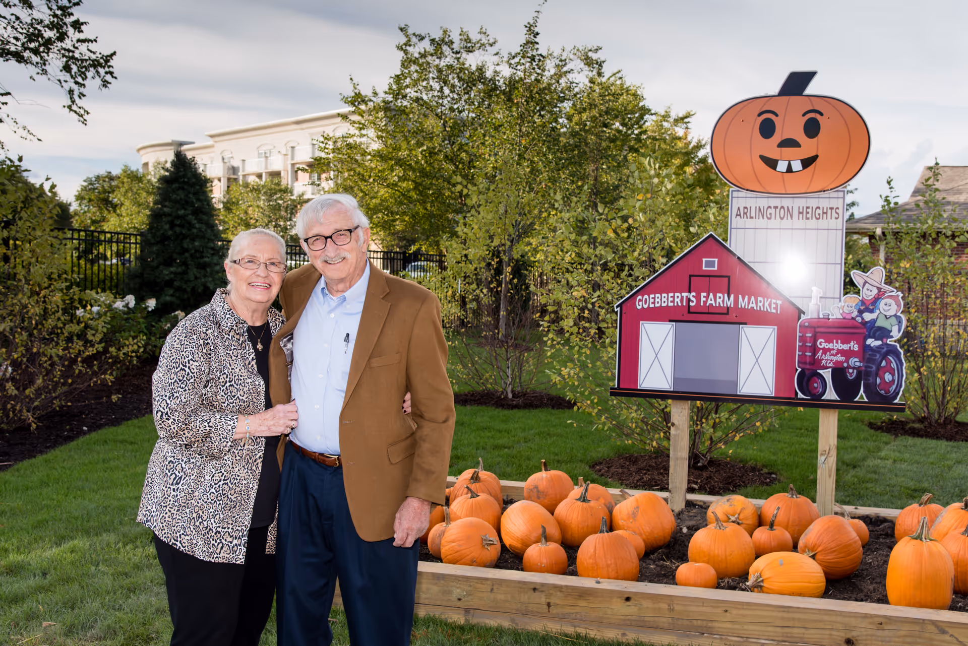 An elderly couple standing outdoors in a garden area with green grass and trees. They are smiling and posing together near a raised wooden planter filled with pumpkins. Behind them is a colorful sign featuring a red barn labeled 'Goebbert's Farm Market' and a large cartoon pumpkin with the text 'Arlington Heights'.
