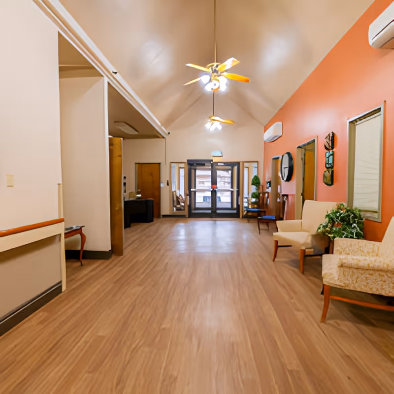 A spacious wood-floored lobby with seating along the right wall, vaulted ceiling with ceiling fans, and double glass entrance doors at the far end.