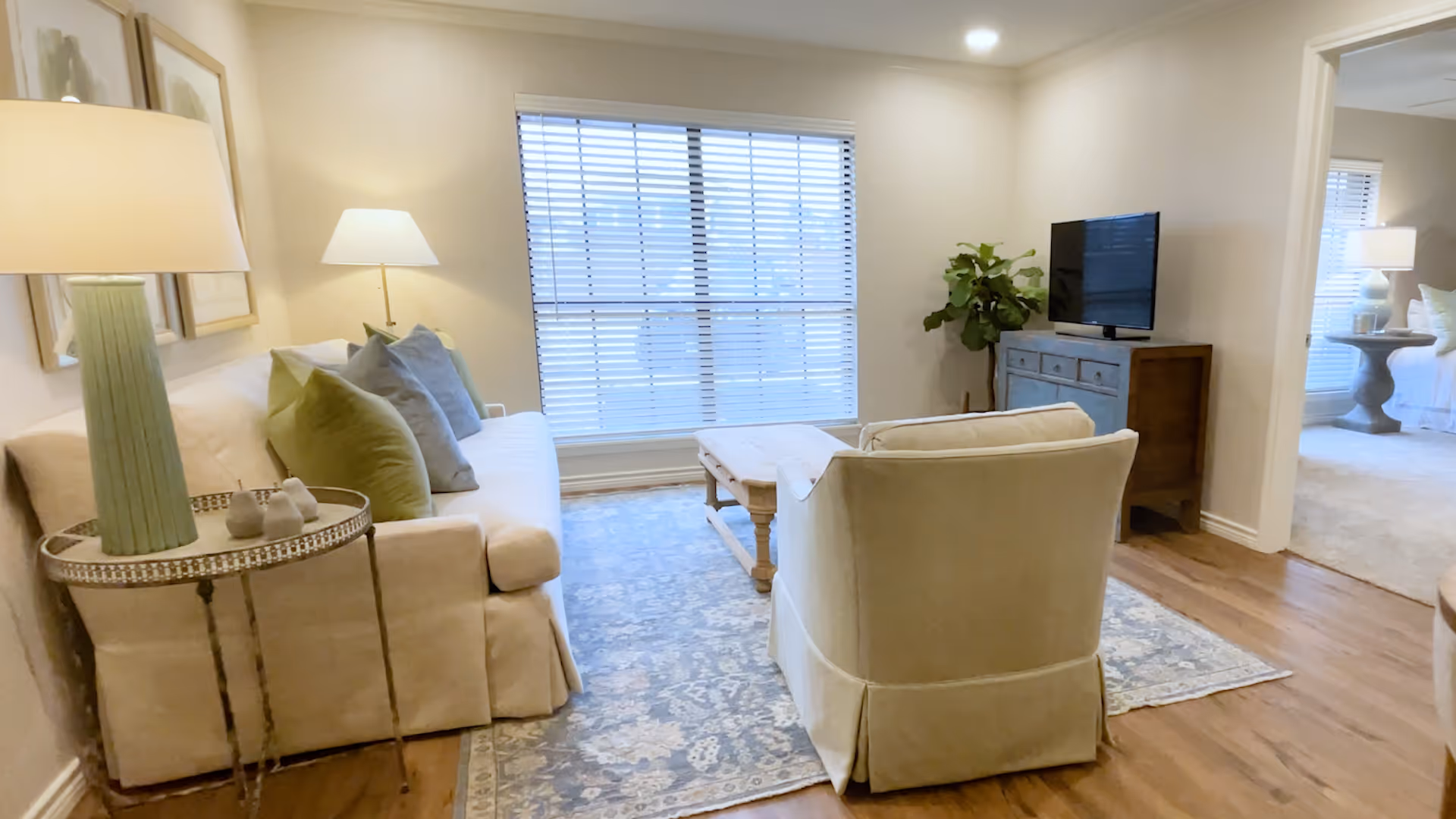 Bright furnished living room with a sofa, armchair, coffee table, TV on a cabinet, and a large window with blinds.