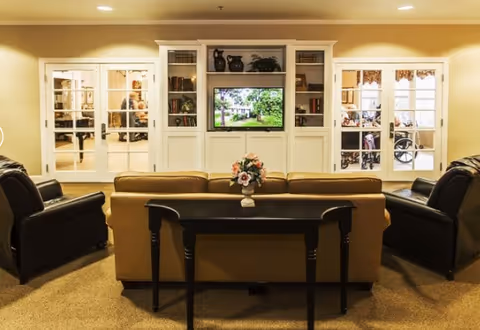 A cozy living room area with a beige sofa and two black armchairs arranged around a black console table with a flower vase. In front of the seating is a white built-in entertainment center with shelves and a TV displaying an outdoor scene. Behind the entertainment center are glass-paneled double doors leading to another room where a person in a wheelchair is visible.