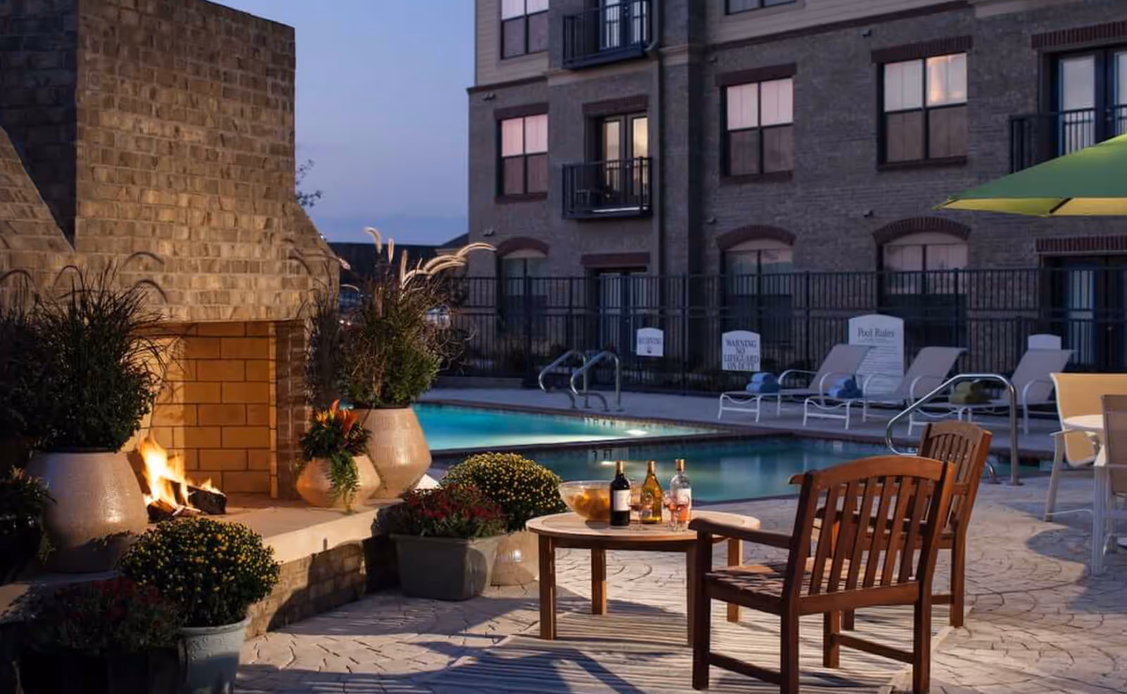 Outdoor patio area at dusk with a lit stone fireplace, potted plants, a wooden table with bottles and a bowl, wooden chairs, a swimming pool with lounge chairs, and a multi-story brick building in the background.