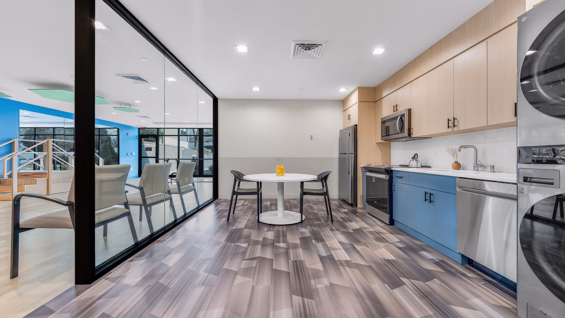 Modern kitchen area in a senior living facility with light wood and blue cabinetry, stainless steel appliances including a refrigerator, microwave, oven, dishwasher, and stacked washer and dryer. A round white table with two black chairs is centered in the room. Large glass wall separates the kitchen from an adjacent seating area with beige chairs and blue walls.