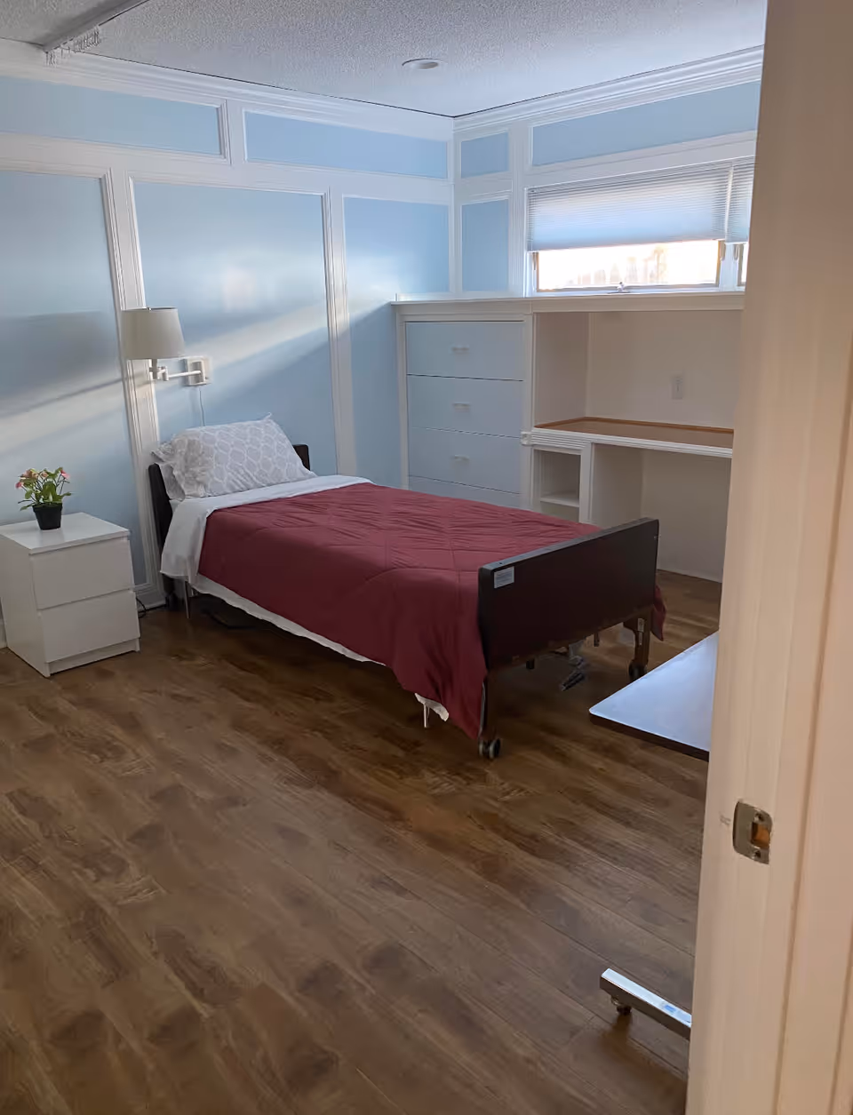 A small bedroom with light blue walls and white trim, featuring a single bed with a maroon quilt and white pillow. There is a white nightstand with a small potted plant, a wall-mounted lamp, a built-in dresser, and a desk area under a window with a partially closed blind. The floor is wood with a natural finish.