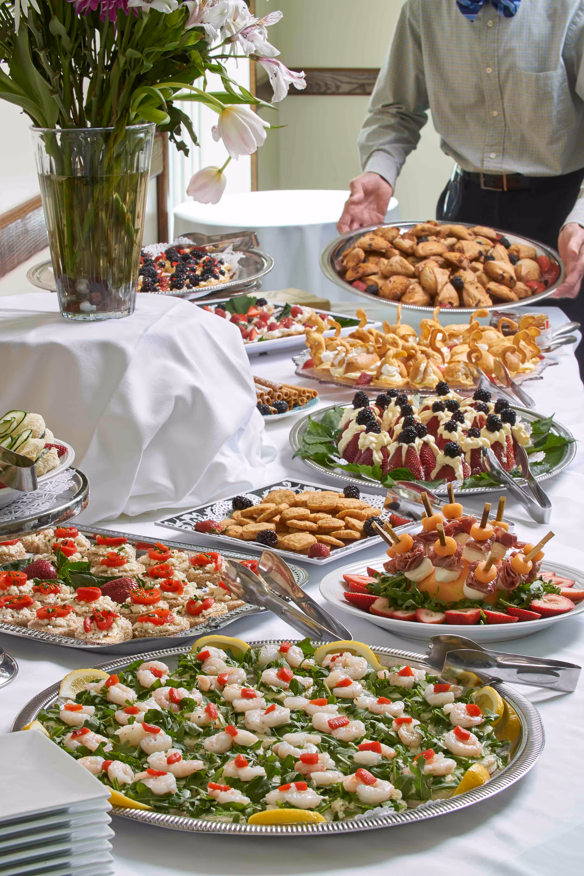 Buffet table with assorted appetizers, pastries and fruit platters, a vase of flowers on the table and a server holding a tray in the background.