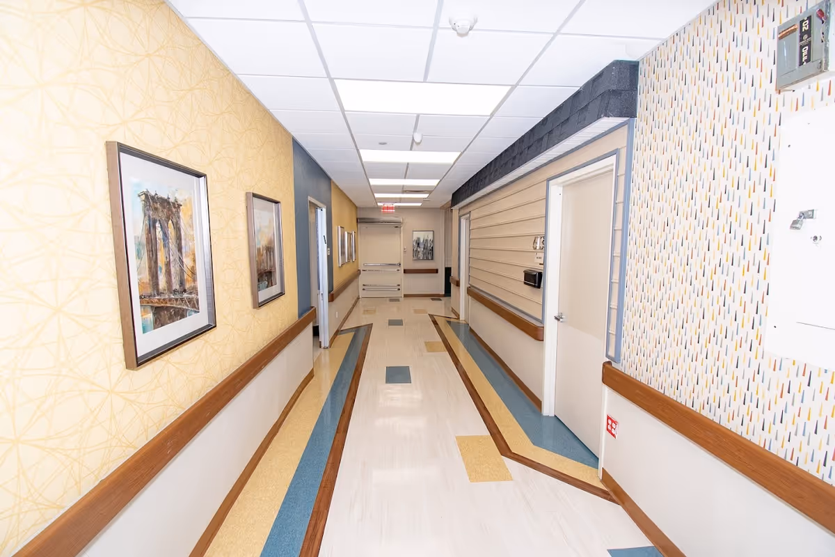 Well-lit interior hallway of a senior living facility with framed artwork, handrails, and patterned flooring.