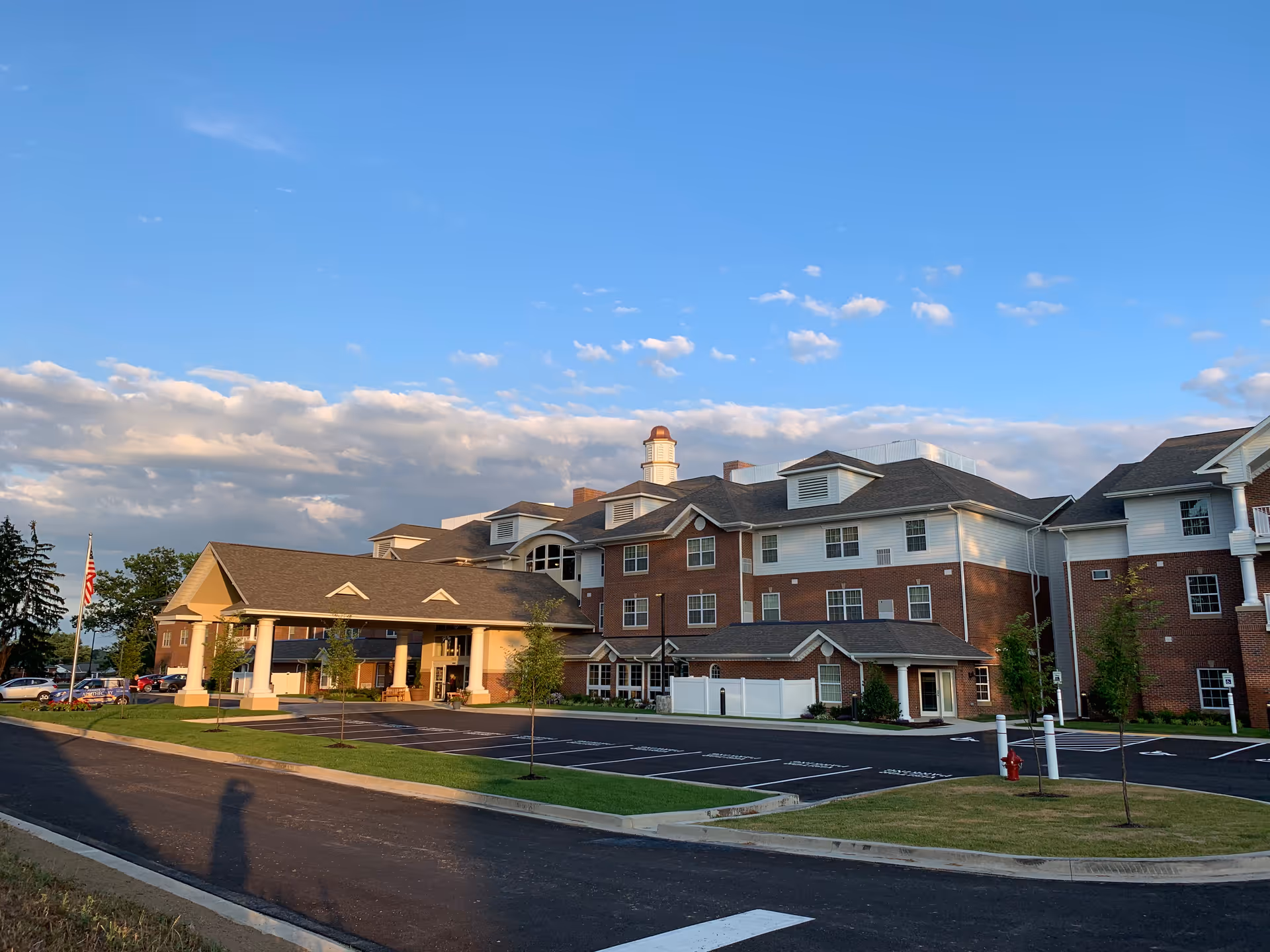 Exterior view of a large, multi-story brick and white siding building with a covered entrance and a parking lot in front, under a partly cloudy blue sky.