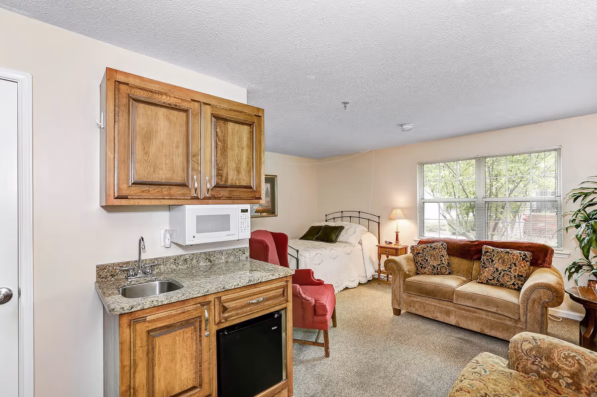 A cozy studio apartment featuring a small kitchenette with wooden cabinets, a granite countertop, a sink, a microwave, and a mini refrigerator. The living area includes a bed with white bedding, two red armchairs, a brown sofa with patterned cushions, a side table with a lamp, and a large window letting in natural light. A potted plant is visible near the window.
