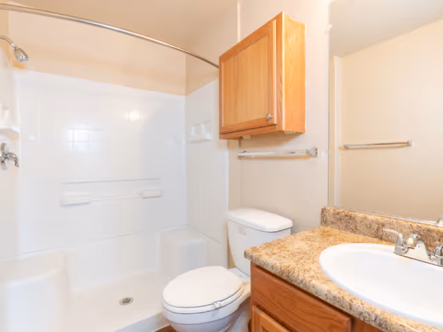 A clean and bright bathroom featuring a white bathtub with a curved shower rod and showerhead, a white toilet, a wooden cabinet mounted on the wall, a granite countertop with a white sink, and a large mirror above the sink.