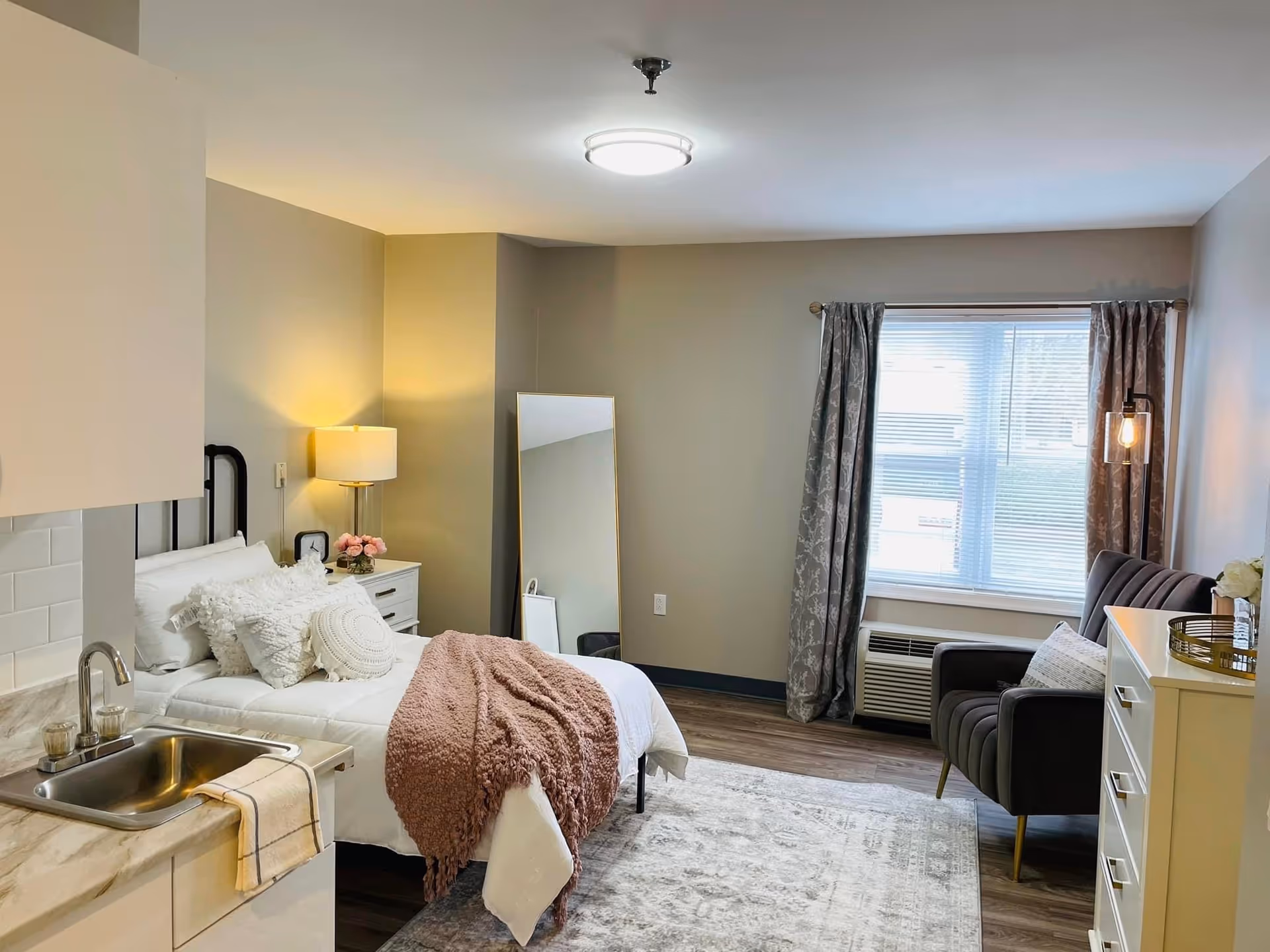 A cozy assisted living bedroom featuring a bed with white bedding and decorative pillows, a pink throw blanket, a bedside table with a lamp and flowers, a full-length mirror leaning against the wall, a window with patterned curtains, a dark upholstered armchair, a white dresser with decorative items, and a small kitchenette area with a sink and countertop.