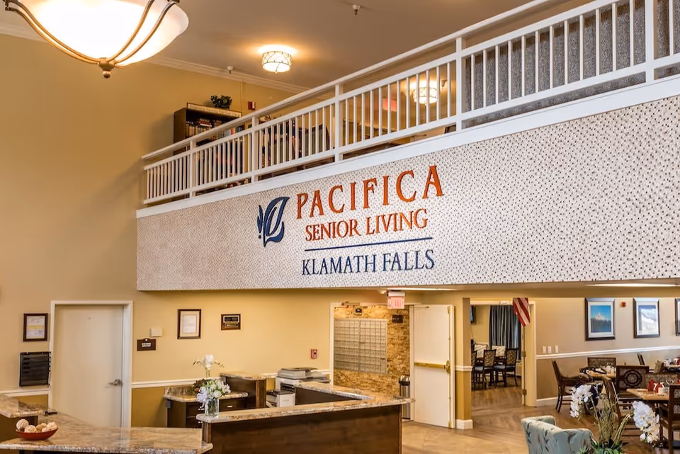 Interior view of the reception area at Pacifica Senior Living Klamath Falls, featuring a front desk with flowers, a sign with the facility name on the wall above, and a glimpse into a dining area with tables and chairs.
