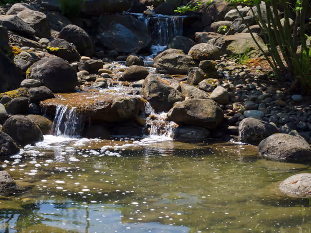 A small naturalistic waterfall flowing over rocks into a clear pond surrounded by stones and greenery.