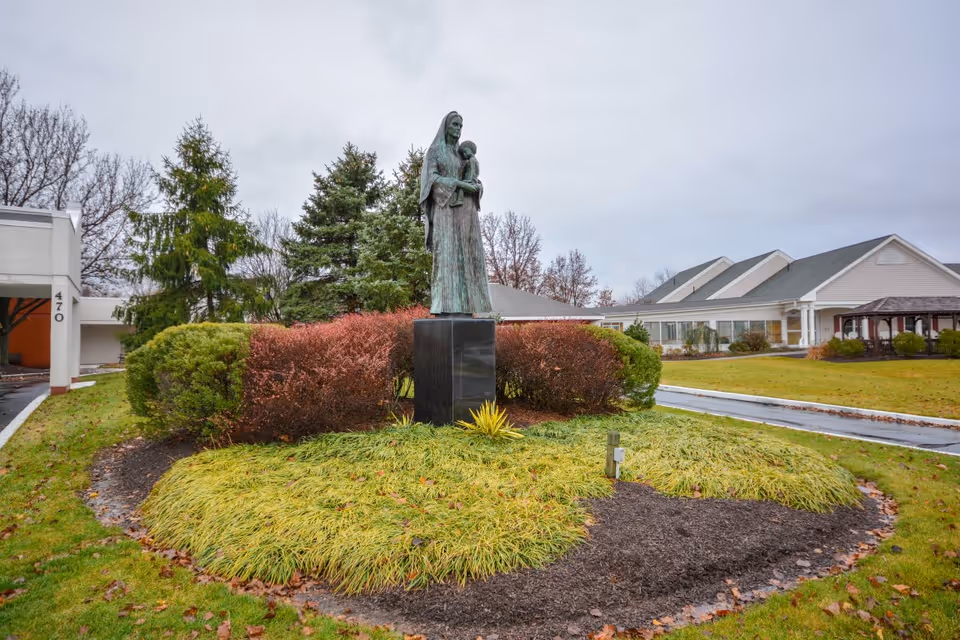 Bronze statue of a woman holding a child in a landscaped circular bed in front of a single-story rehabilitation facility under an overcast sky.