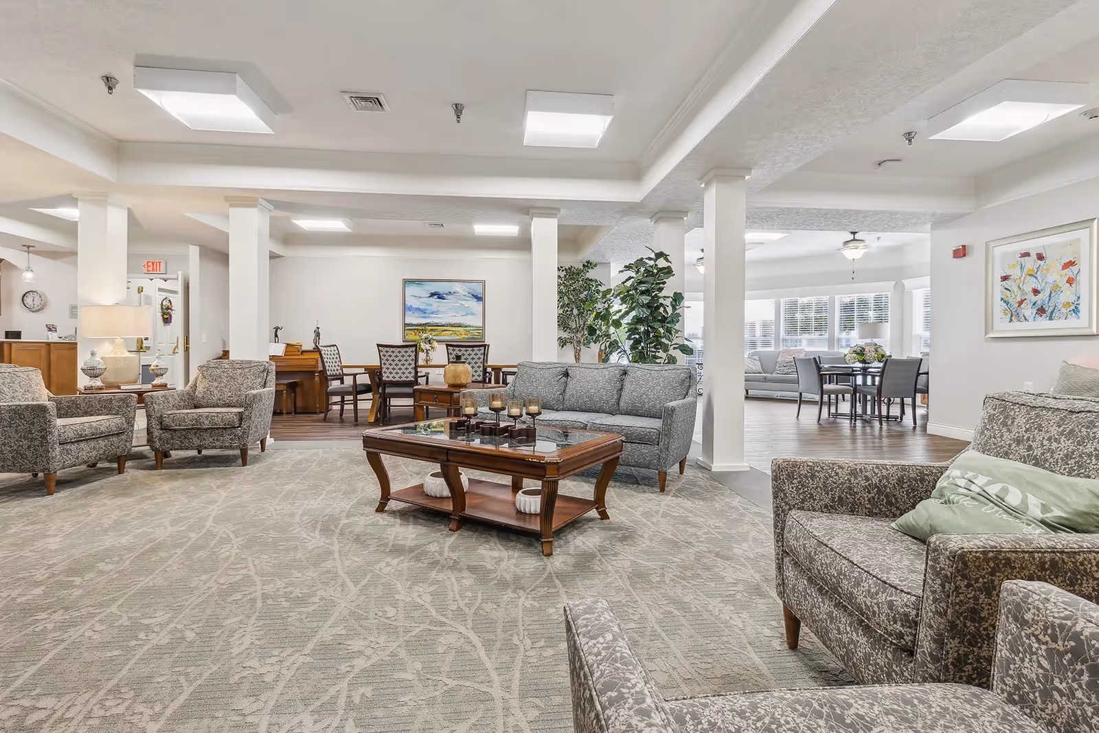 A spacious and well-lit senior living facility common area with multiple seating arrangements including patterned armchairs and sofas around a wooden coffee table. The room features a light-colored carpet with a subtle branch pattern, white walls, and several columns. In the background, there is a dining area with tables and chairs, large windows letting in natural light, and framed artwork on the walls.