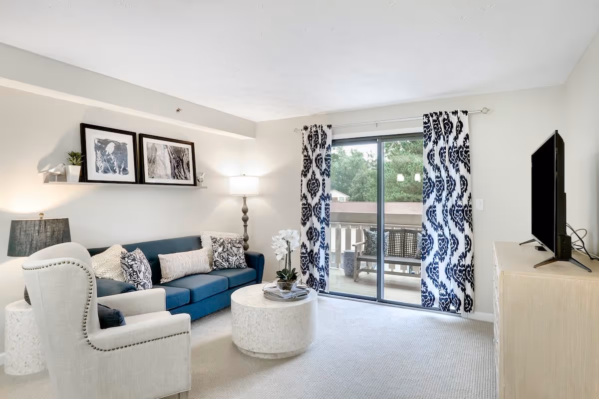 A bright living room with a blue sofa adorned with patterned cushions, a beige armchair, a round white coffee table with a flower arrangement, a floor lamp, and a TV on a light-colored stand. The room has sliding glass doors with blue and white patterned curtains leading to a balcony with outdoor seating and greenery visible outside.