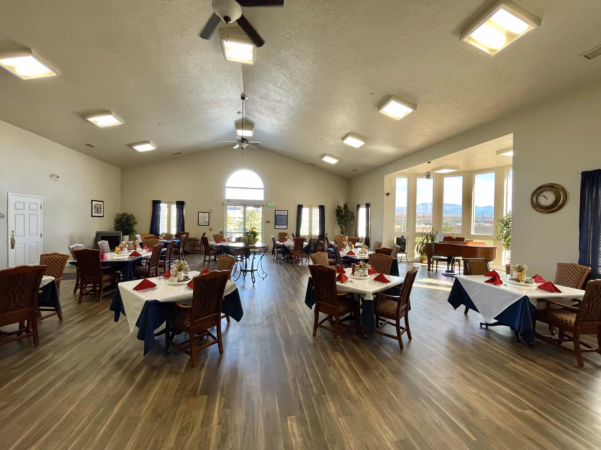 A spacious dining room with multiple tables covered in white and navy blue tablecloths, each set with red folded napkins and condiments. The room has wooden flooring, large windows letting in natural light, ceiling fans, and a grand piano near the windows. There are plants and framed pictures on the walls, creating a welcoming atmosphere.