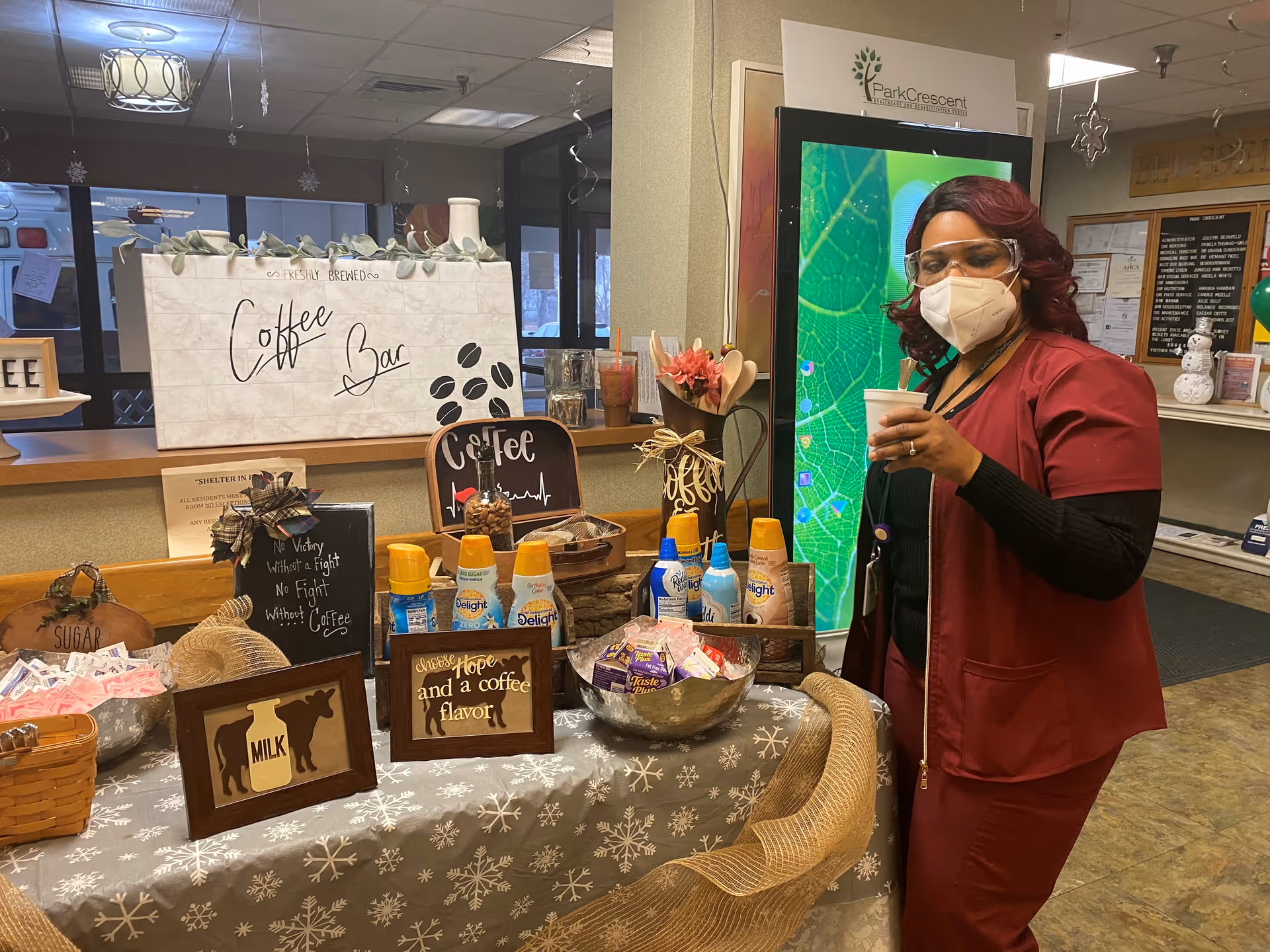 A woman wearing a red uniform, face mask, and protective glasses stands next to a decorated coffee bar table inside a healthcare facility. The table is covered with a snowflake-patterned cloth and displays various coffee creamers, sugar packets, and signs with coffee-related quotes. Behind the table is a sign that reads 'Coffee Bar' and a digital screen with a green leaf image. The setting appears to be a reception or common area of the Park Crescent Healthcare and Rehabilitation Center.