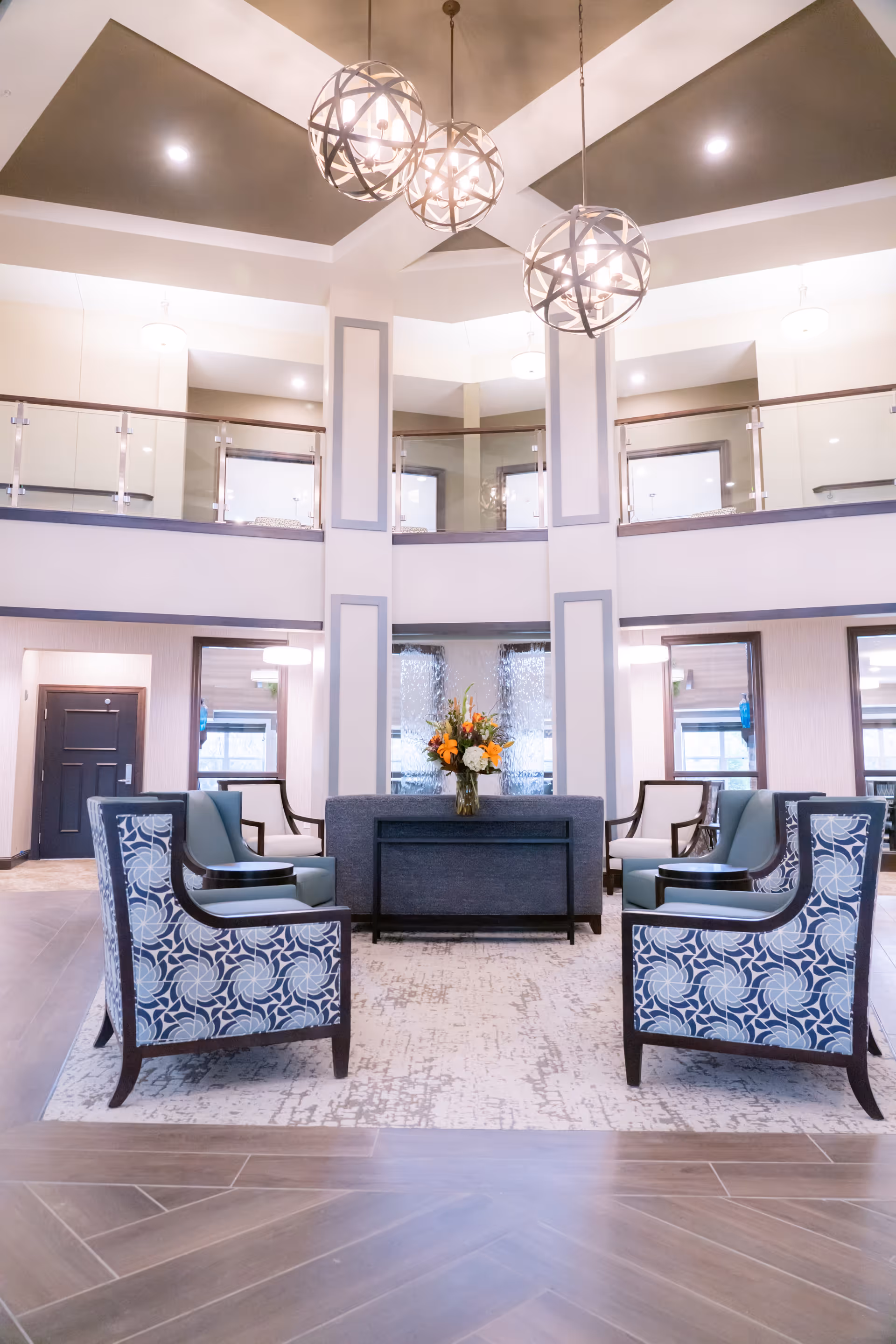 A spacious and well-lit senior living facility common area with a high ceiling and modern spherical pendant lights. The seating area includes patterned armchairs and a sofa arranged around a central table with a vase of flowers. The upper level features glass railings overlooking the area below.