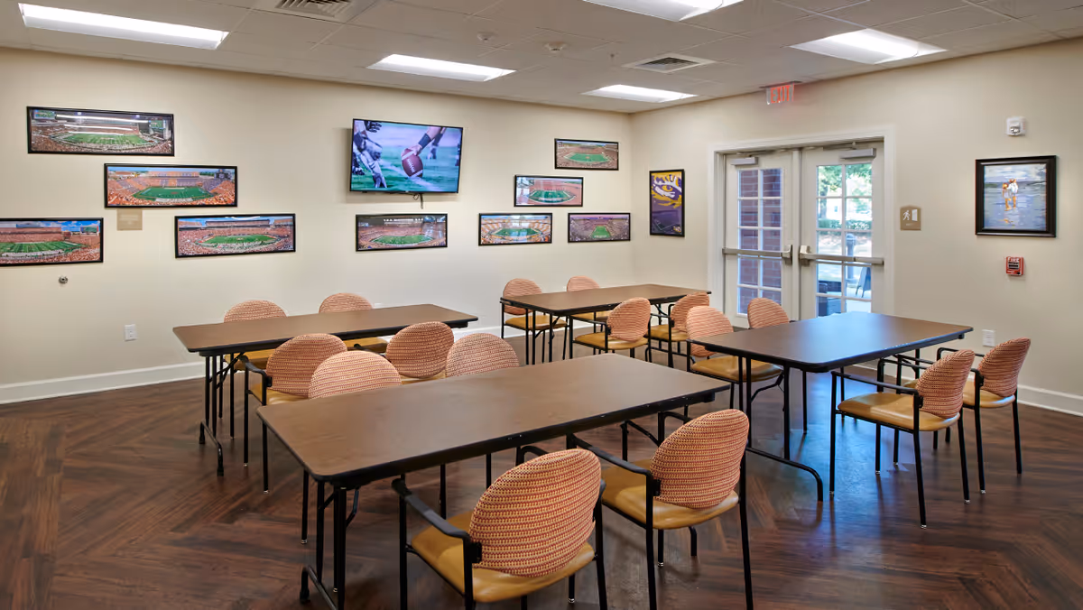 A well-lit room with several rectangular tables and cushioned chairs arranged in rows. The walls are decorated with framed pictures, mostly of sports fields and a football player. There is a double glass door leading outside, and the floor has a wood pattern.