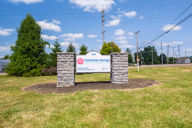 Stone-framed sign reading 'Coshocton Springs' on a grassy lawn with trees and a road in the background.