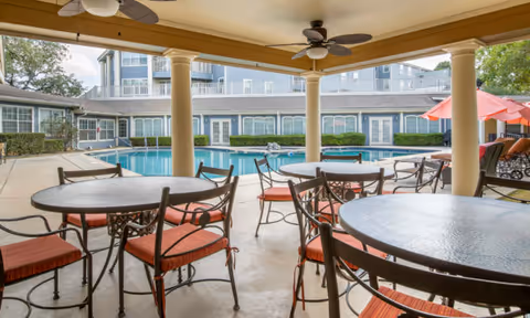 Covered outdoor patio with round tables and red-cushioned chairs overlooking a swimming pool and surrounding community buildings.