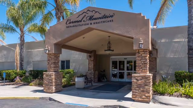 Entrance of Carmel Mountain Rehabilitation and Healthcare Center featuring a covered driveway with stone pillars, palm trees, and a clear blue sky.
