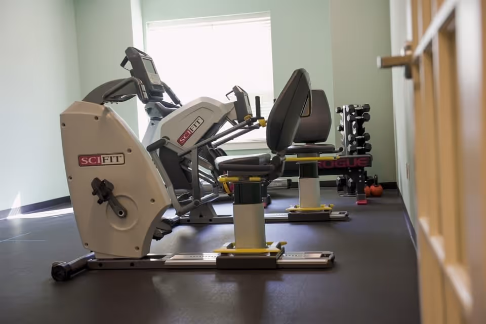 A small exercise room with two SciFit recumbent exercise bikes positioned side by side near a window. In the background, there is a rack of dumbbells and some kettlebells on the floor. The room has light green walls and a dark floor.