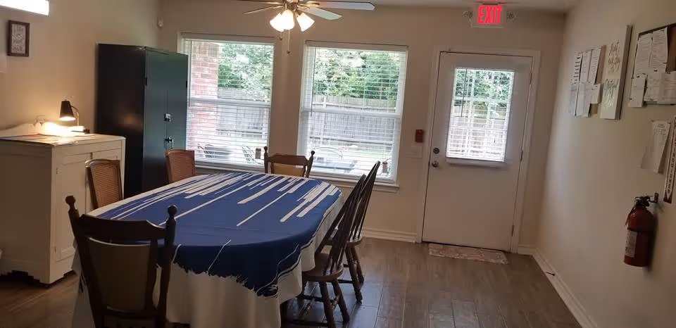 Communal dining room with a rectangular table covered by a blue tablecloth, wooden chairs, windows and a door with an exit sign.