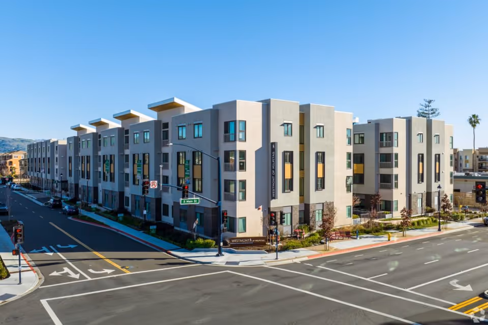 Modern multi-story residential building at a street intersection under a clear blue sky. The building has large windows and a sign that reads 'Westmont of Milpitas'. Traffic lights and street signs are visible, including one for S Main St.