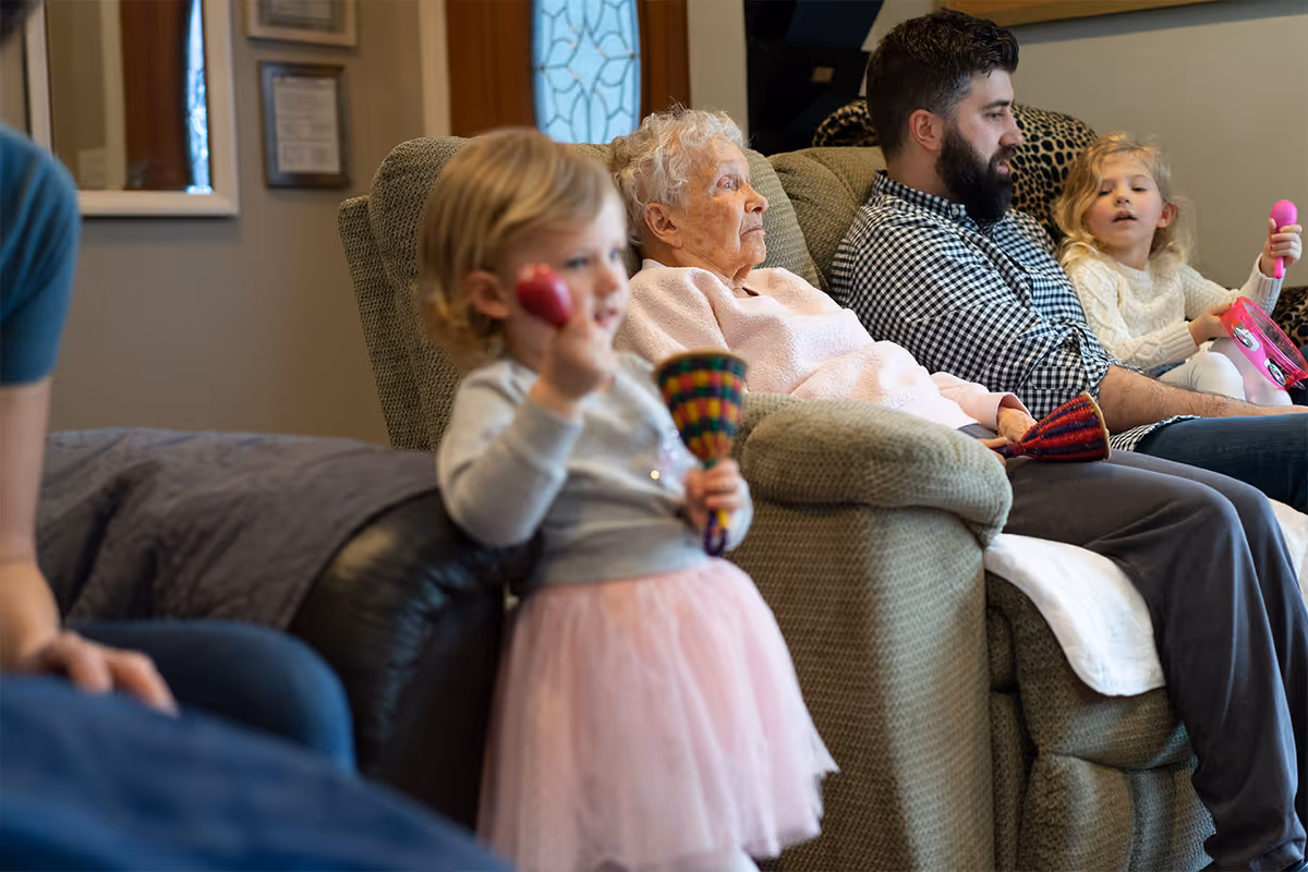 An elderly woman sitting on a beige recliner chair in a living room with a man and two young children nearby. One child is standing in the foreground holding colorful maracas, while the other child is sitting next to the man holding a pink toy. The setting appears warm and home-like.