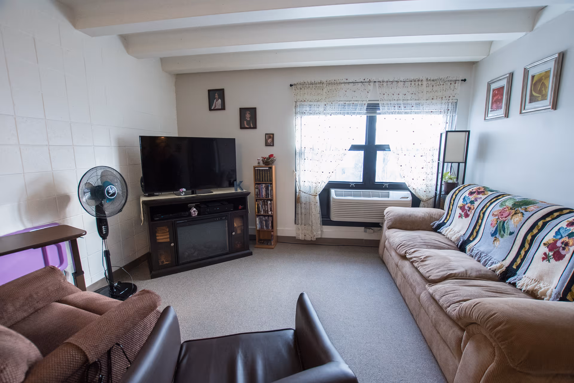 A cozy living room with a large beige couch covered with floral-patterned blankets, a brown recliner chair, a standing fan, a flat-screen TV on a dark wooden stand, and a window with sheer curtains letting in natural light. The walls have a few framed pictures and the floor is carpeted.