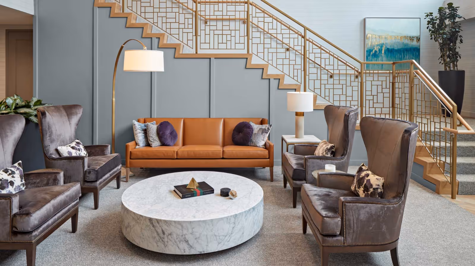 A stylish living room area featuring a tan leather sofa with decorative pillows, four dark gray upholstered armchairs with patterned cushions, a large round white marble coffee table with books and decorative items, a floor lamp, a table lamp on a side table, and a staircase with a gold geometric railing. A large plant and a blue and gold abstract painting are visible in the background.